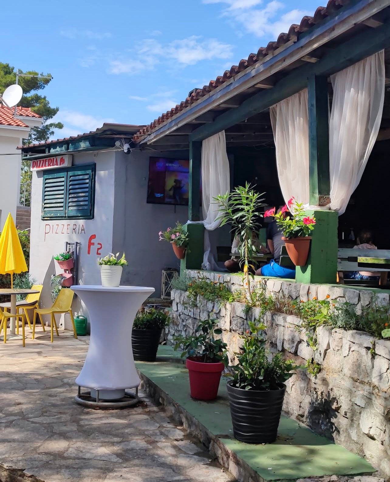 Ulaz i terasa Pizzerije F2 na Jadriji: natpis PIZZERIA F2 na bijelom zidu, kameni zid s cvijećem u teglama, žute stolice pod suncobranom i plavo nebo